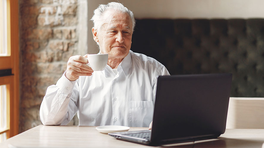 Older man sitting at a table in front of an open laptop.