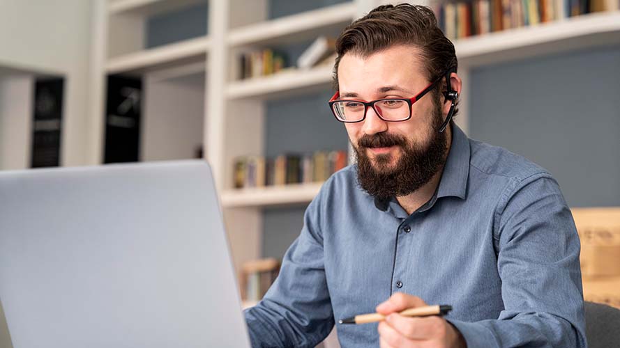 Teacher working at a laptop in a library. He's wearing headphones and holding a pen