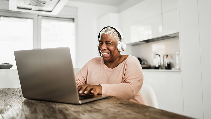 Smiling lady, wearing headphones uses a laptop in her kitchen