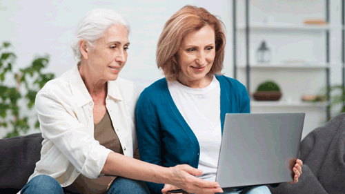 Two mature women, sat on a sofa, looking at a laptop screen.
