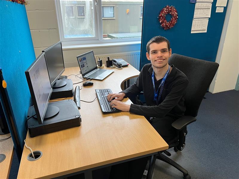 Harley sits at his desk, working on his computer in the Dolphin office. He is wearing a dark jumper and is smiling.